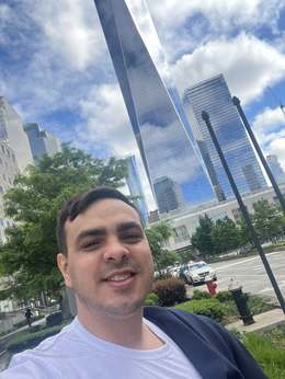 Jônatas Vasconcelos sorrindo em frente aos prédios do World Trade Center em Nova York